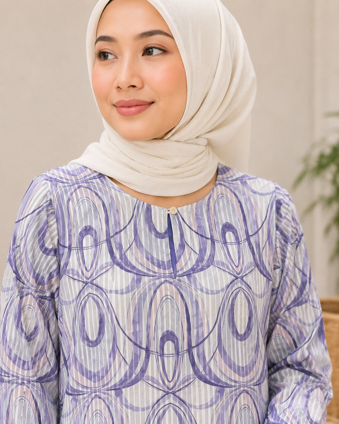 A woman wearing a light beige hijab and a long-sleeve blouse with a purple and white abstract circular pattern, shown in a softly lit indoor setting.