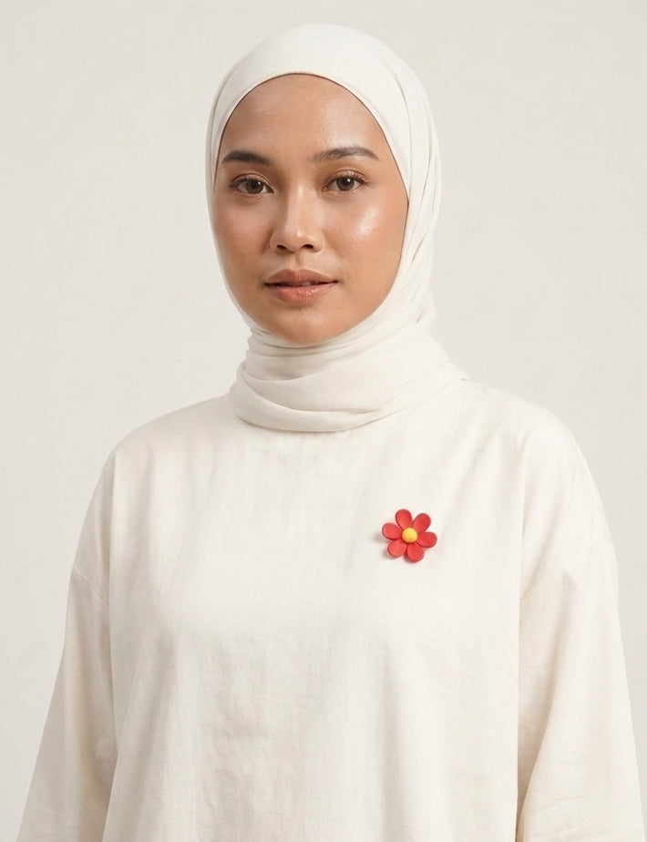 A woman wearing a cream-colored hijab and matching long-sleeve top, featuring a small red flower brooch on the chest, set against a plain light background.