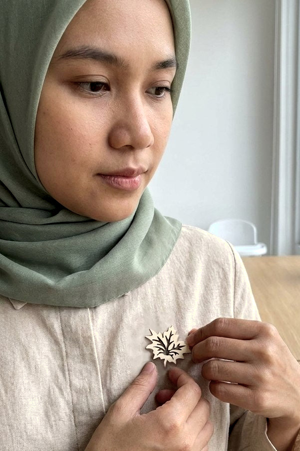 A close-up lifestyle shot of a woman wearing a sage green hijab and a light beige linen top, adjusting a small, laser-cut wooden leaf-shaped brooch on her chest.