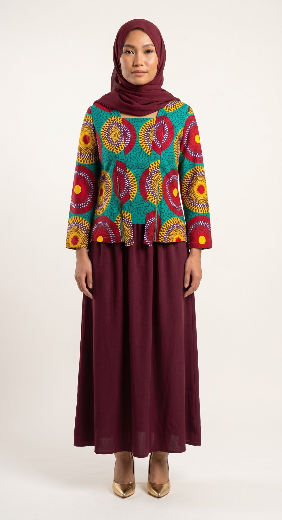 A woman modeling a long-sleeve blouse with a vibrant multicolor circular pattern on a teal background, paired with a solid burgundy maxi skirt and matching hijab, against a plain light background.