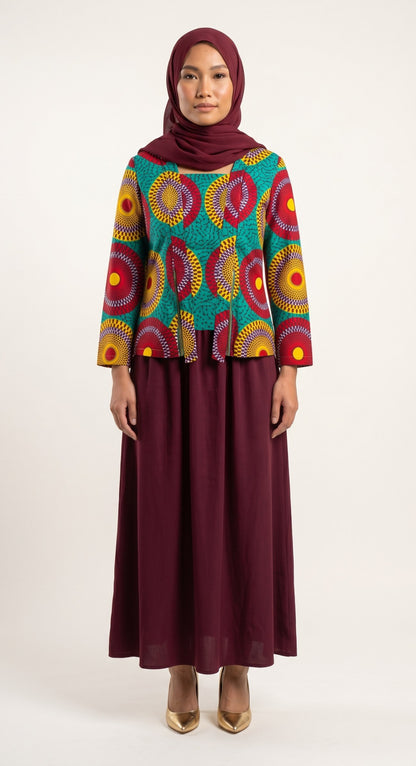 A woman modeling a long-sleeve blouse with a vibrant multicolor circular pattern on a teal background, paired with a solid burgundy maxi skirt and matching hijab, against a plain light background.