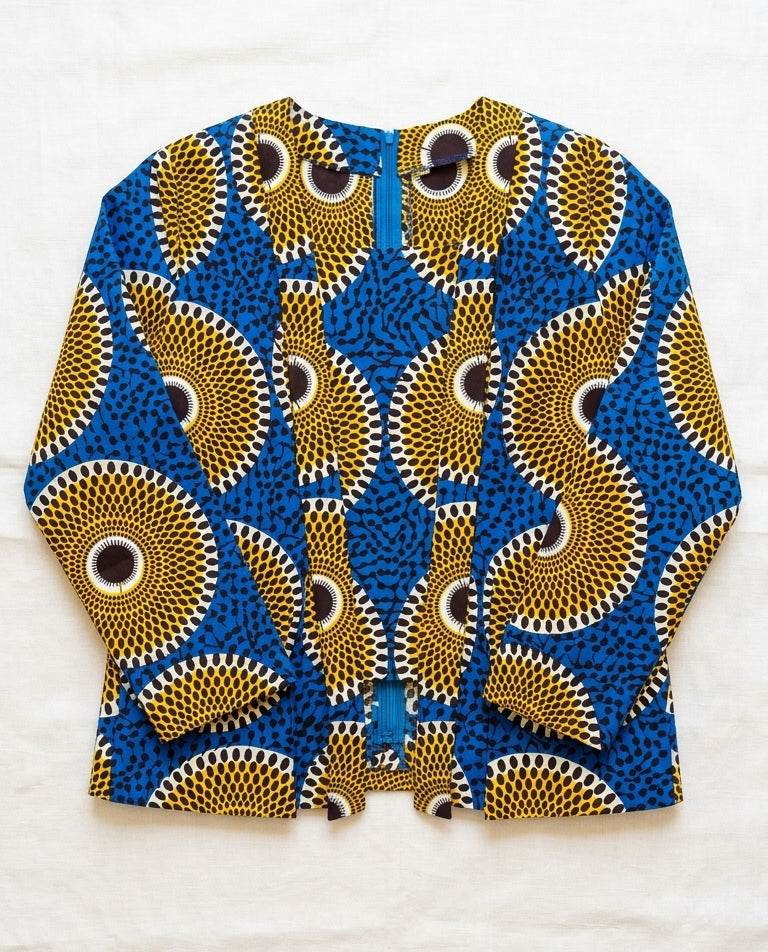 A long-sleeve cardigan featuring a vibrant blue and mustard yellow African wax print pattern with circular eye-like motifs, laid flat on a white textured background.