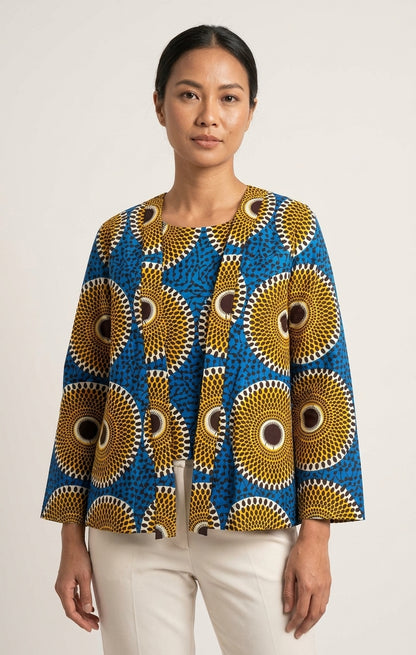 A woman modeling a long-sleeve blouse with a vibrant blue, gold, and white African wax print pattern, paired with cream-colored pants, against a plain light gray background.