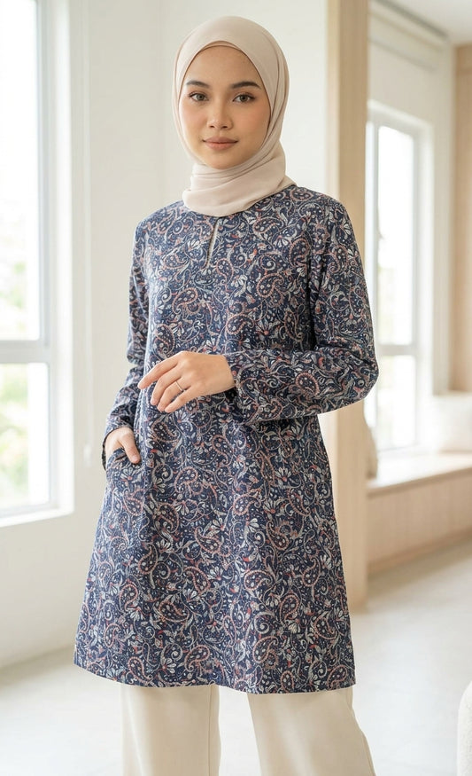 A woman wearing a long-sleeved tunic with a blue, white, and red paisley pattern, paired with a beige hijab and cream pants, standing in a bright, modern room.