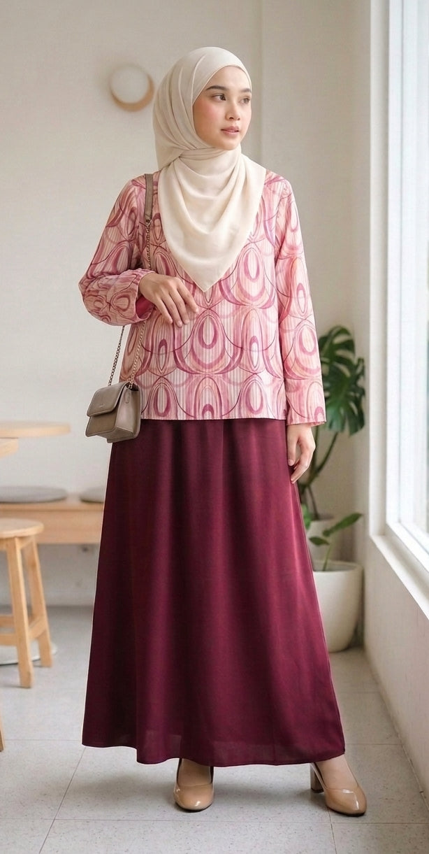 A woman wearing a long-sleeve pink and white patterned blouse, a deep burgundy maxi skirt, and a cream hijab, carrying a small taupe shoulder bag in a bright indoor setting.
