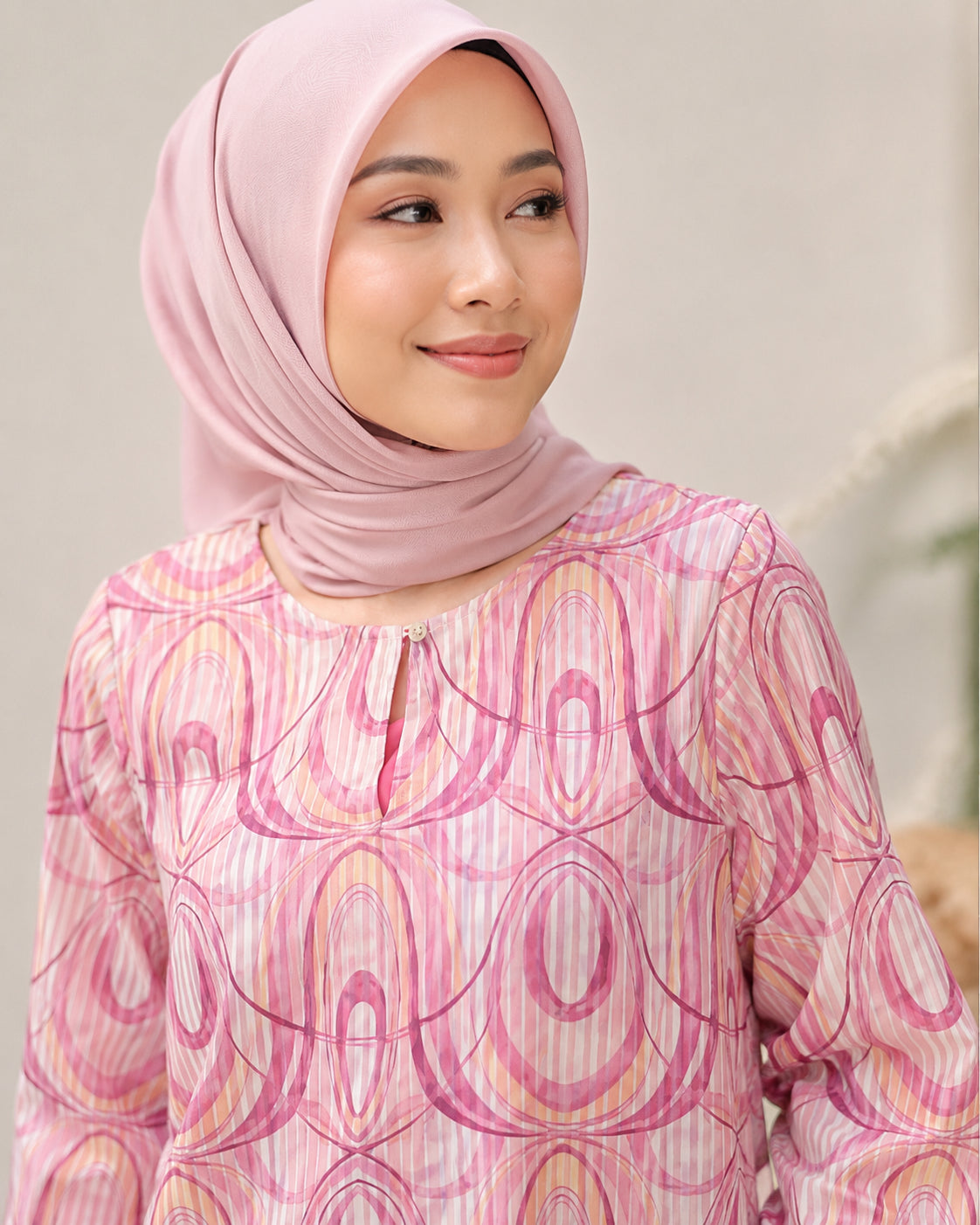 A woman wearing a light pink hijab and a matching pink long-sleeve blouse with a pink, purple, and yellow abstract oval pattern, shown against a soft beige background.