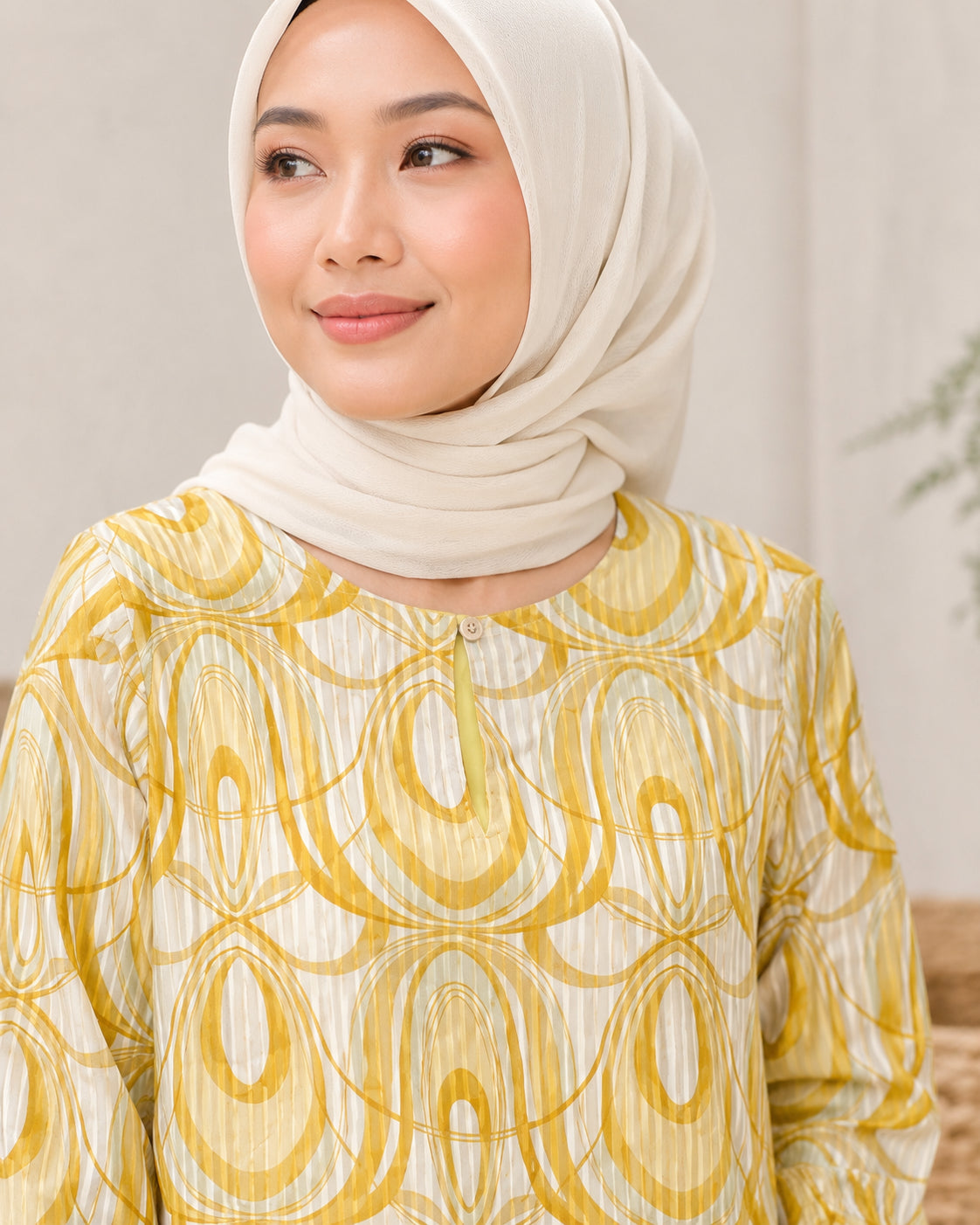 A woman wearing a cream-colored hijab and a matching cream blouse with a yellow and white abstract oval pattern, shown in a close-up portrait.