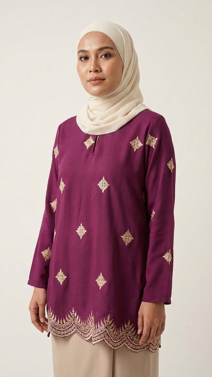 A woman wearing a long-sleeve purple tunic with gold geometric embroidery and a scalloped hem, paired with a cream hijab and light beige trousers, photographed against a plain light background.