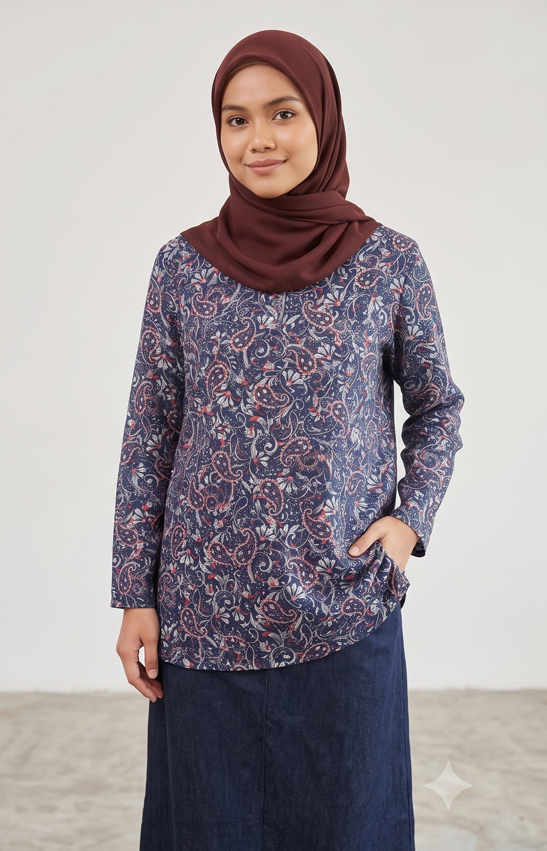 A woman wearing a long-sleeved navy blue tunic top with a red and white paisley pattern, paired with a dark navy skirt and a maroon hijab, standing against a light gray studio background.