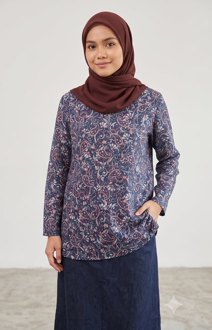 A woman wearing a long-sleeved navy blue tunic top with a red and white paisley pattern, paired with a dark navy skirt and a maroon hijab, standing against a light gray studio background.