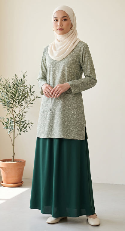 A woman wearing a light sage green long-sleeve tunic and a dark teal maxi skirt, paired with a cream hijab and beige shoes, standing in a bright indoor setting next to a potted plant.