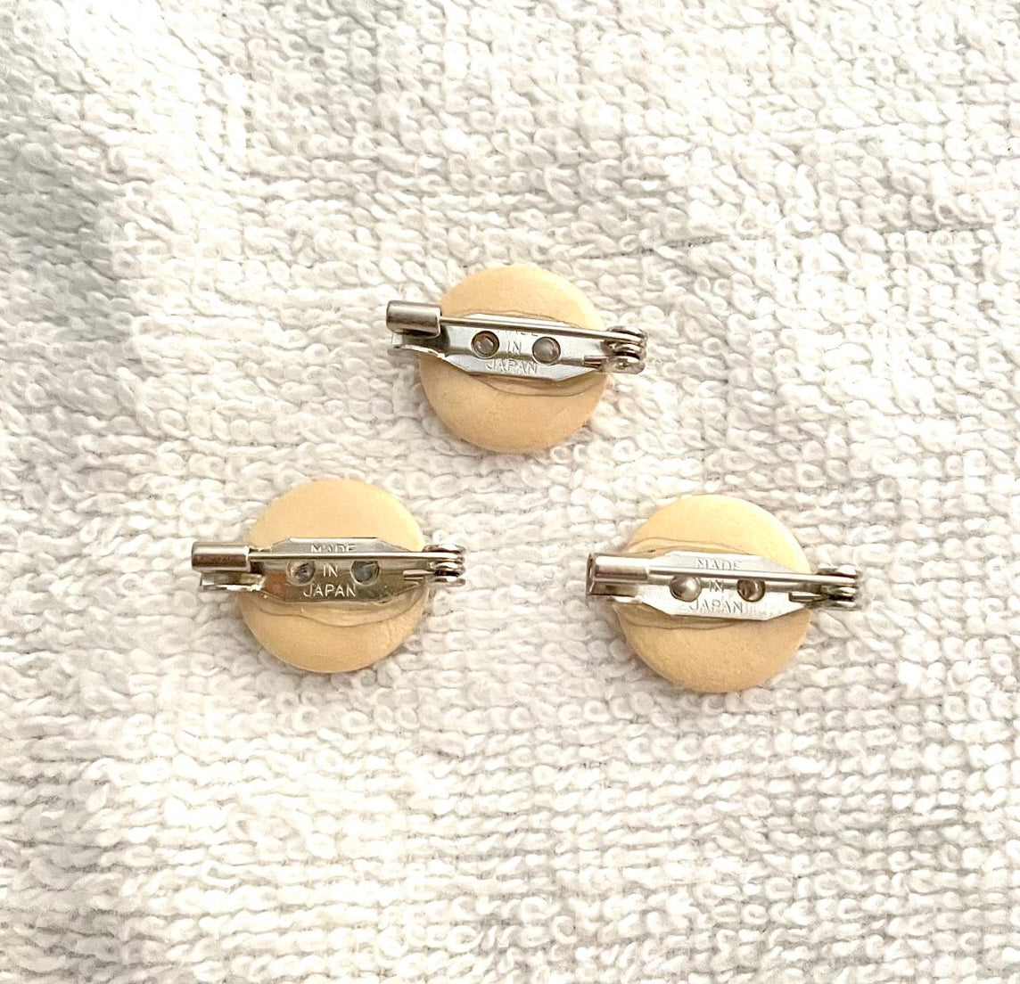 Three round beige pin-back buttons with silver metal safety bar clasp backs, displayed on a textured white fabric background.