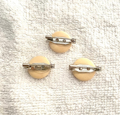 Three round beige pin-back buttons with silver metal safety bar clasp backs, displayed on a textured white fabric background.