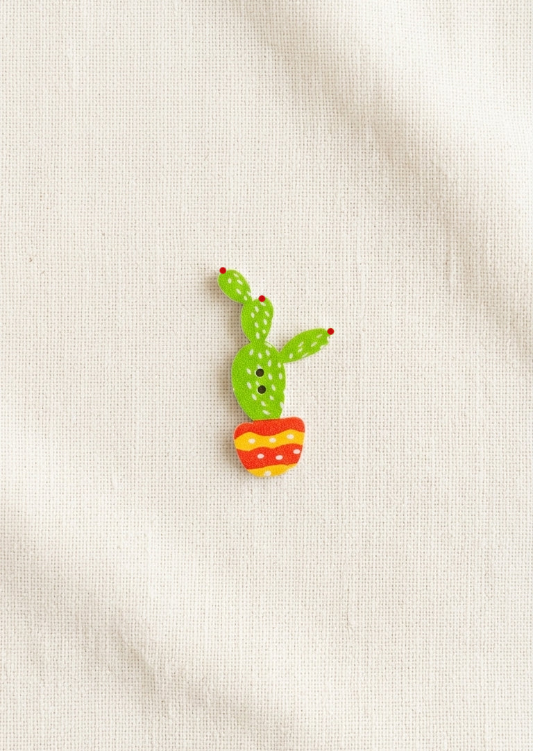 A small wooden pin shaped like a green cactus in a red and yellow striped pot, placed on a textured off-white fabric background.