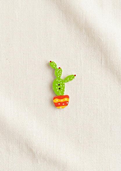 A small wooden pin shaped like a green cactus in a red and yellow striped pot, placed on a textured off-white fabric background.