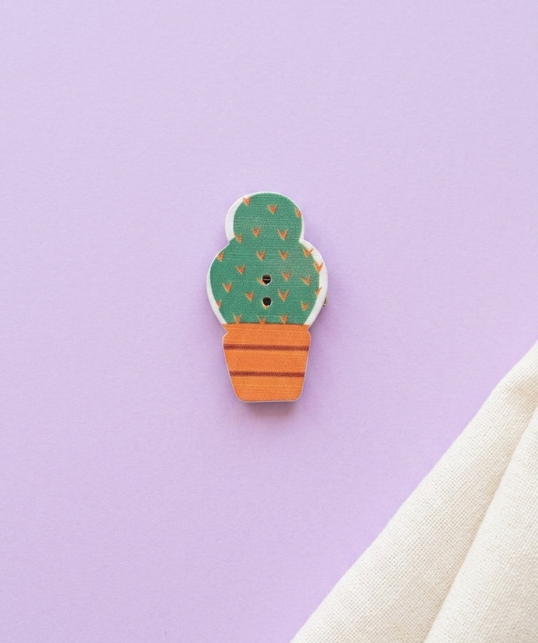 A small cactus-shaped button brooch with a green body, orange pot, and tiny orange leaf details, placed on a light purple background with a folded cream fabric at the bottom right.