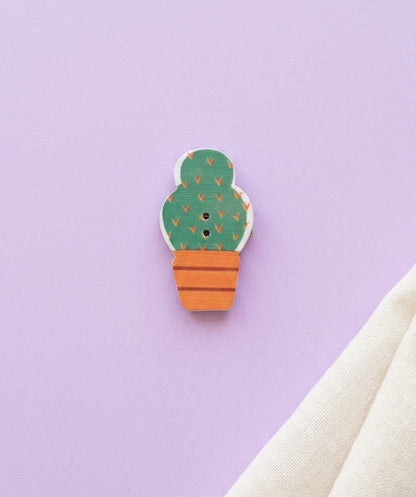 A small cactus-shaped button brooch with a green body, orange pot, and tiny orange leaf details, placed on a light purple background with a folded cream fabric at the bottom right.