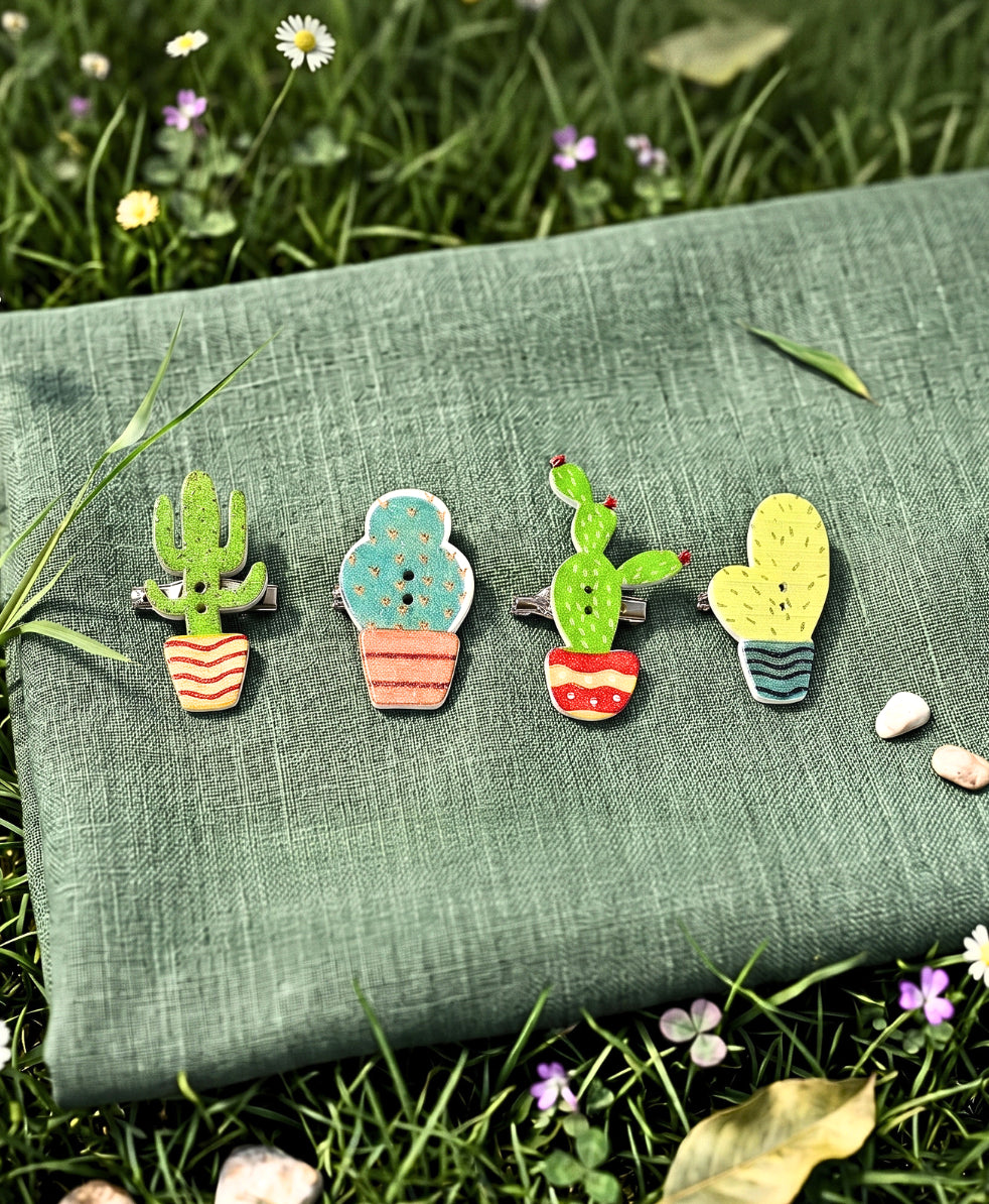 Four colorful wooden cactus hair pins in different pot designs, arranged on a green linen cloth with grass and small wildflowers in the background.