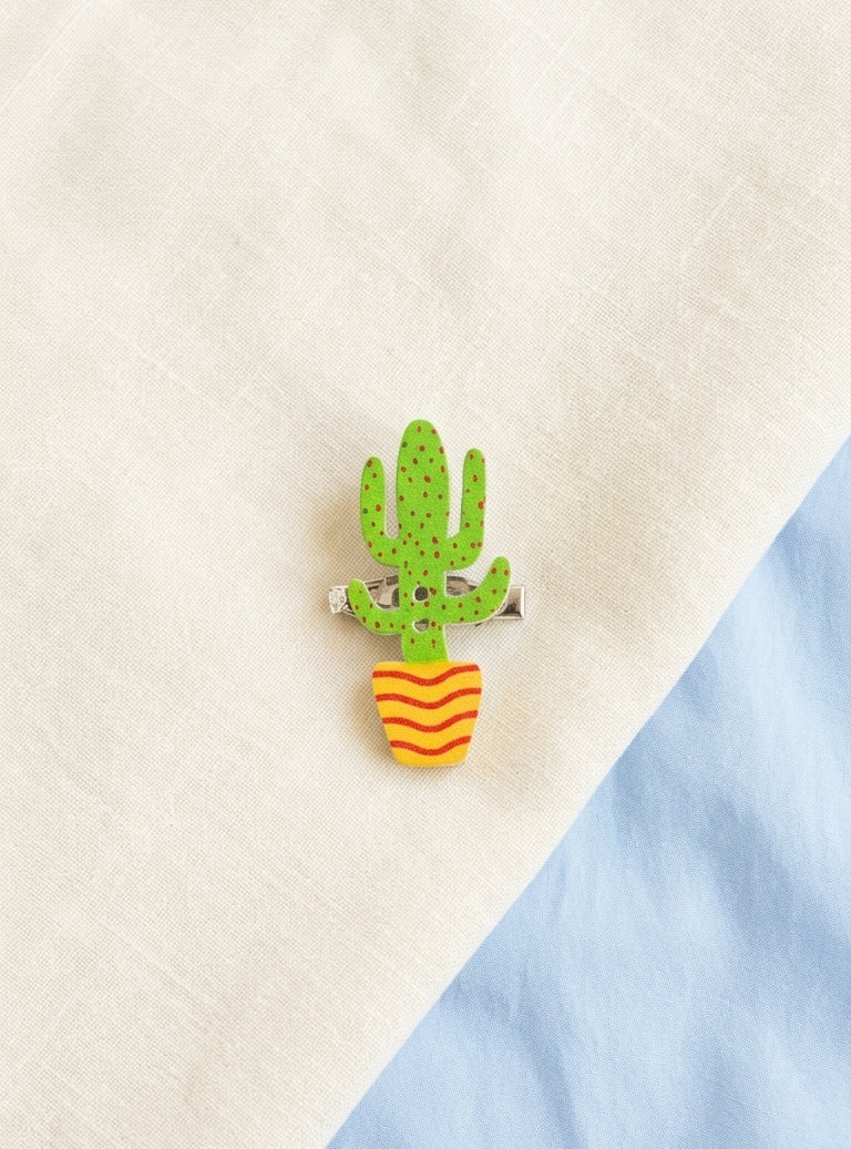 A small wooden pin shaped like a green cactus in a yellow-orange striped pot, photographed on a cream fabric background with a light blue fabric section at the lower right.