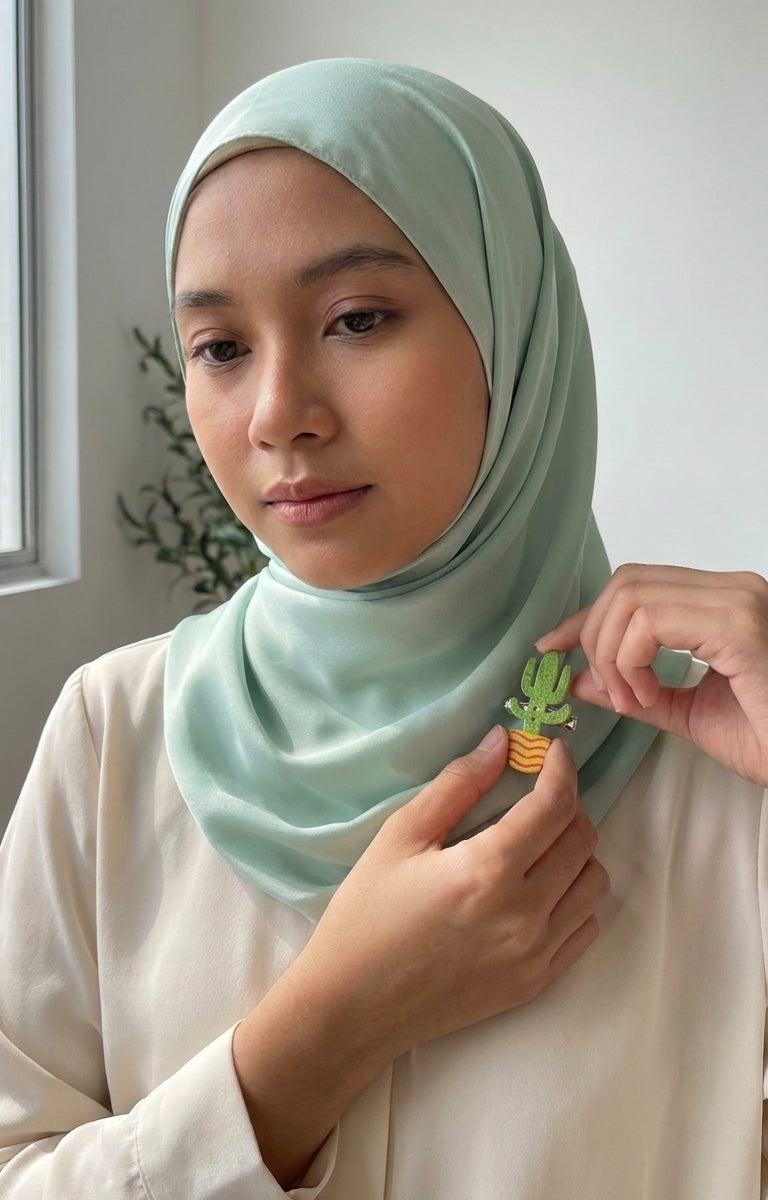 A woman wearing a sage green hijab pinches a small green cactus brooch from another person's hand, set against a soft, neutral indoor background.