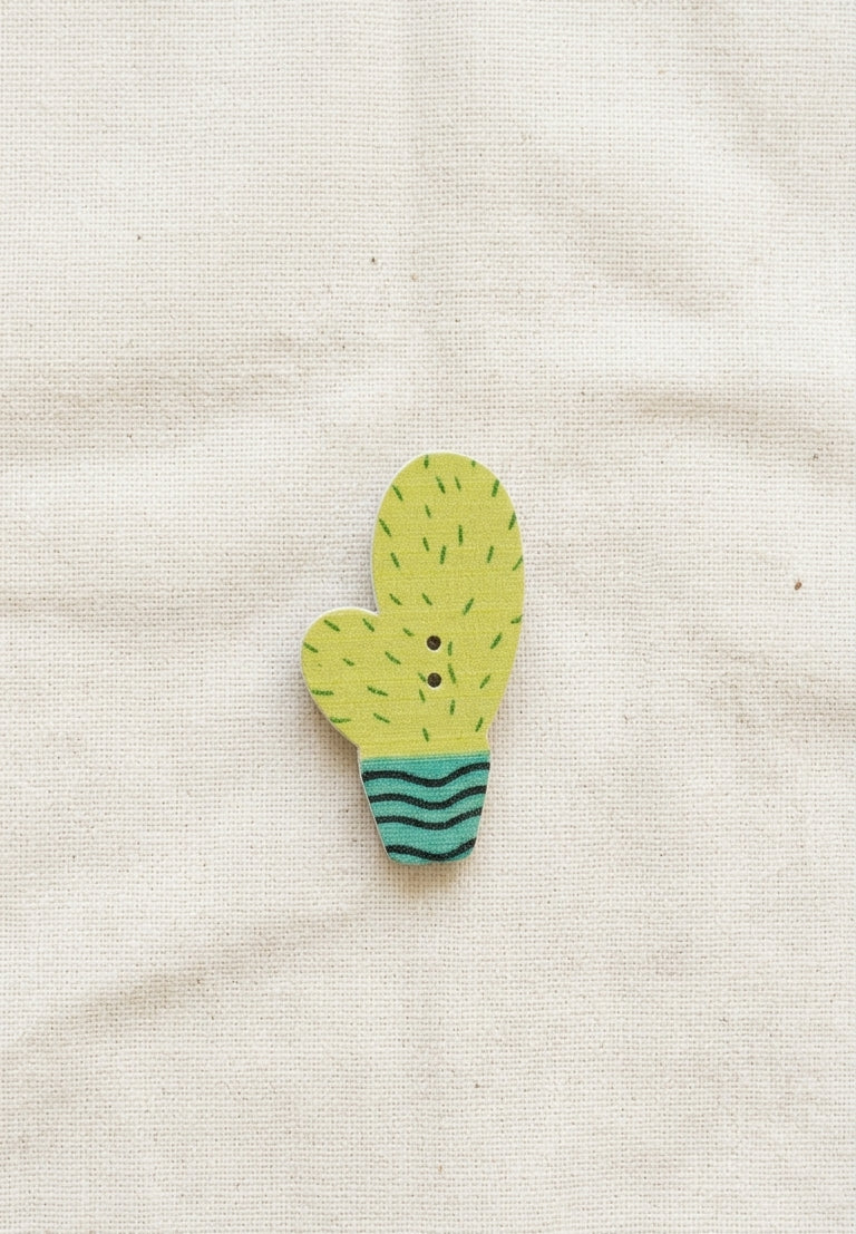 A small cactus-shaped wooden brooch with a yellow-green body, black spines, and a teal striped base, displayed on a textured beige fabric background.