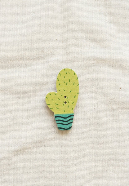 A small cactus-shaped wooden brooch with a yellow-green body, black spines, and a teal striped base, displayed on a textured beige fabric background.