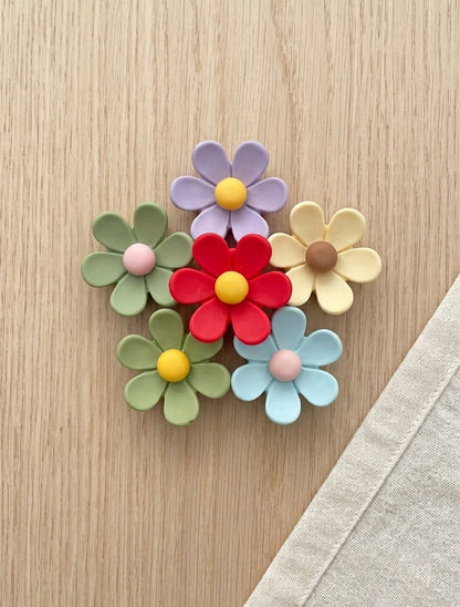 Five colorful flower-shaped brooches arranged on a light wood surface, with a beige fabric corner visible in the lower right. The flowers are in pastel green, lavender, red, pale yellow, and light blue, with yellow, pink, and brown centers.