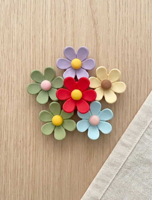 Five colorful flower-shaped brooches arranged on a light wood surface, with a beige fabric corner visible in the lower right. The flowers are in pastel green, lavender, red, pale yellow, and light blue, with yellow, pink, and brown centers.