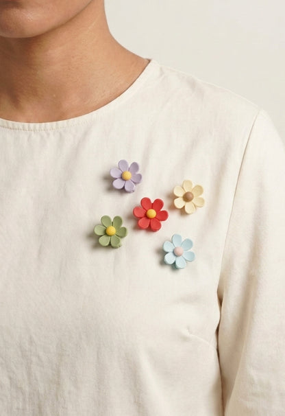 A close-up of a person wearing a cream-colored long-sleeve top with five colorful flower-shaped brooches attached near the left chest. The flowers are in pastel shades of purple, green, red, yellow, and light blue.
