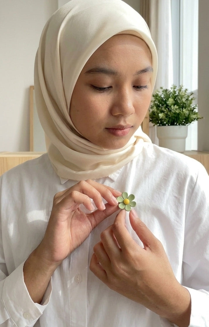 A woman wearing a cream-colored hijab and white shirt holds a small green flower with yellow accents near her chest.