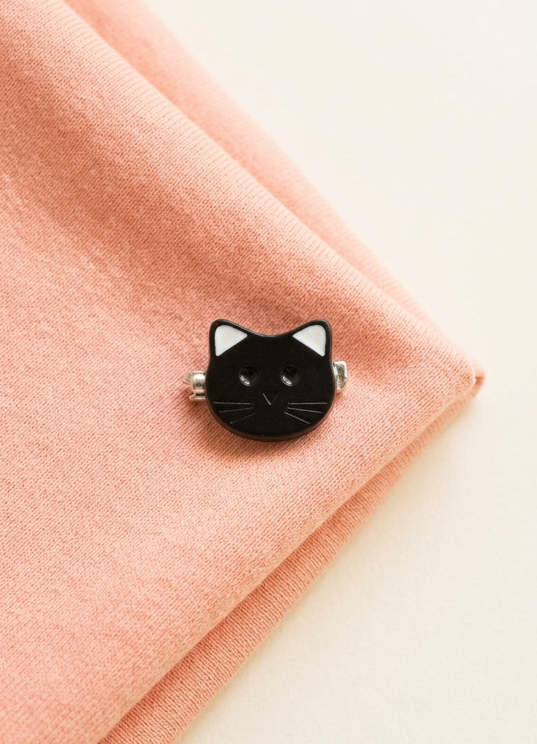 A small black cat-face pin with white inner ears and silver metal sides, placed on peach-pink textured fabric against a light cream background.