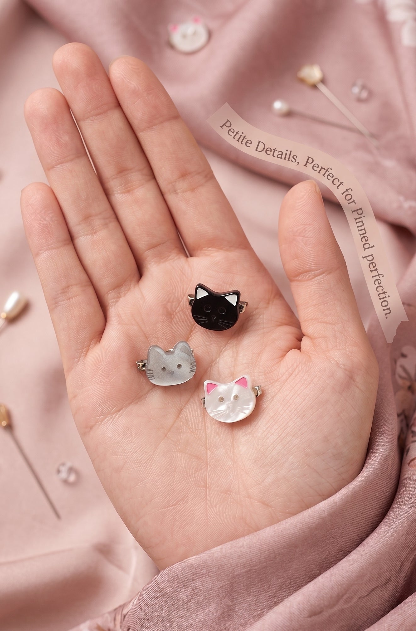 a palm showing 3 mini cat faced mini brooches on a pink background