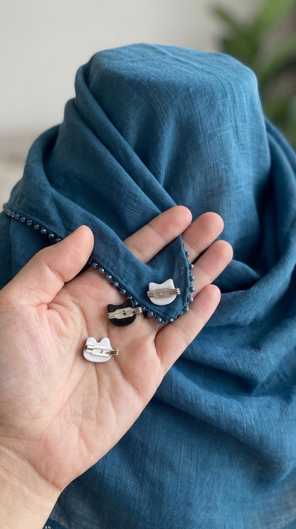a palm showing the back side of the cat faced mini brooches to show the brooch pins with a background of a mannequin of greenish shawl