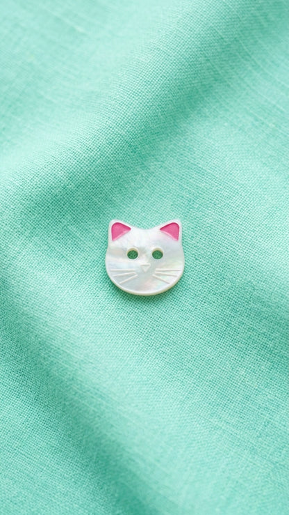 A small white cat-face button with pink ears with 2 holes, placed on a textured mint green fabric background.
