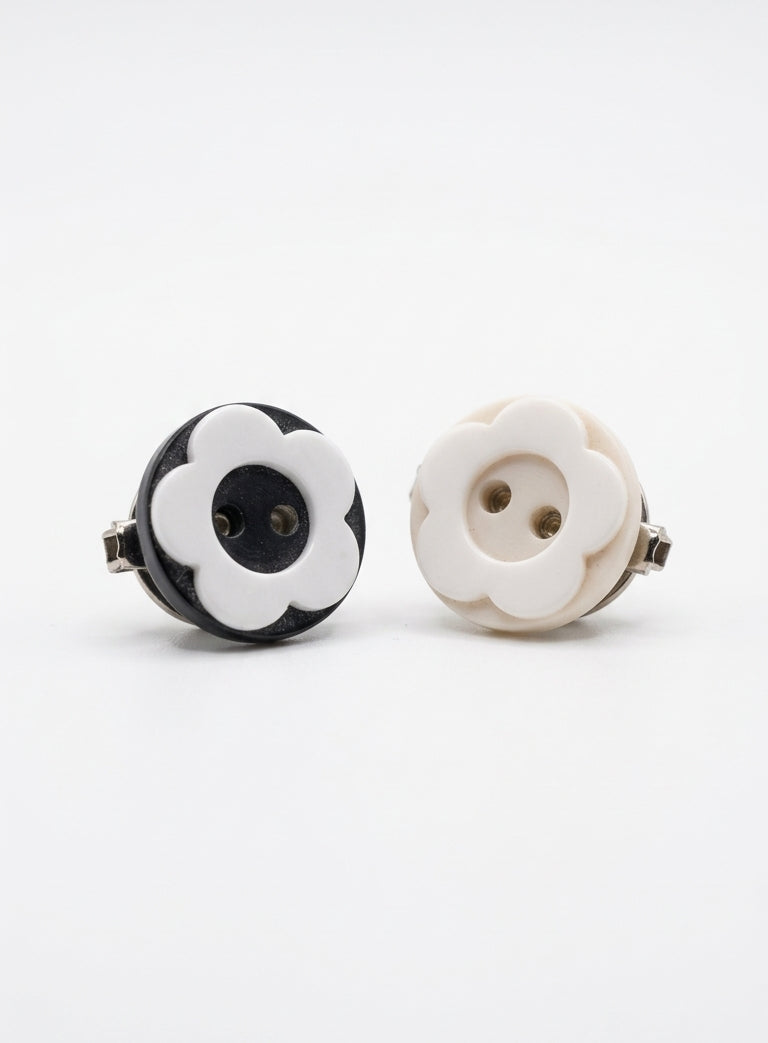 Two round flower-shaped earrings with metal backs, shown on a clean white background. One earring is black and white, and the other is cream and white.