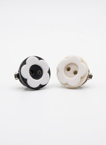 Two round flower-shaped earrings with metal backs, shown on a clean white background. One earring is black and white, and the other is cream and white.