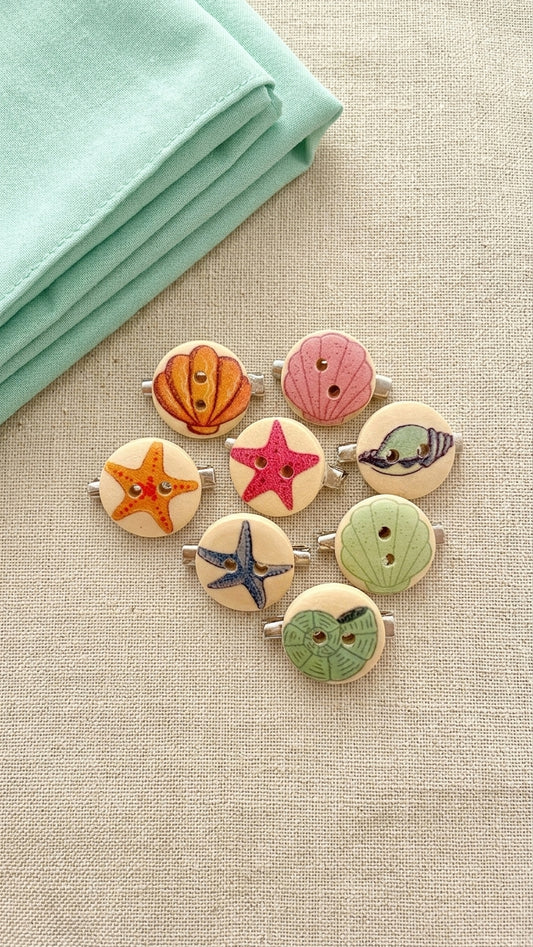 A set of nine round wooden button mini brooches featuring hand-painted nautical designs like starfish, shells, and fish, arranged on a beige linen fabric next to a folded mint green cloth.