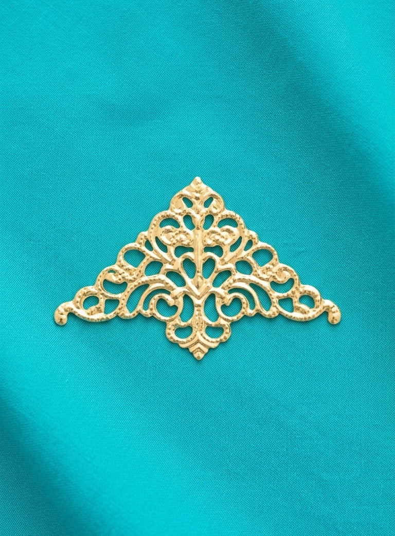 An ornate gold metal filigree applique with a symmetrical, decorative scrollwork pattern, displayed on a textured turquoise blue fabric background.