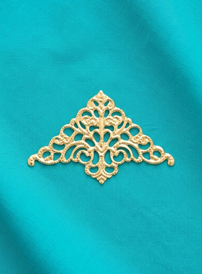 An ornate gold metal filigree applique with a symmetrical, decorative scrollwork pattern, displayed on a textured turquoise blue fabric background.