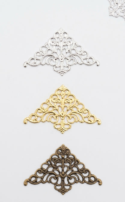 Three ornate filigree corner embellishments in silver, gold, and antique bronze finishes, arranged vertically on a plain white background.