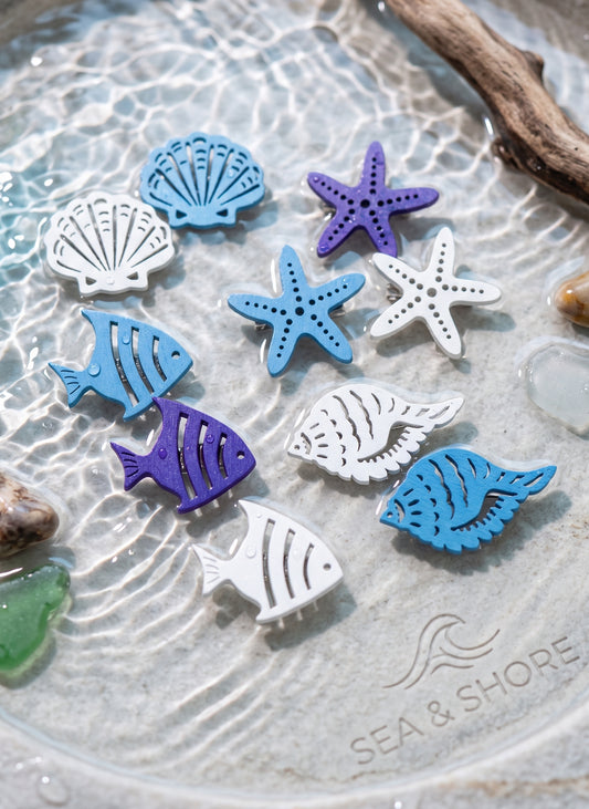 A set of colorful sea-themed wooden brooches, including blue and purple starfish, white and blue seashells, and blue and purple fish, arranged on a textured glass plate with the brand name 'SEA & SHORE' printed on it.