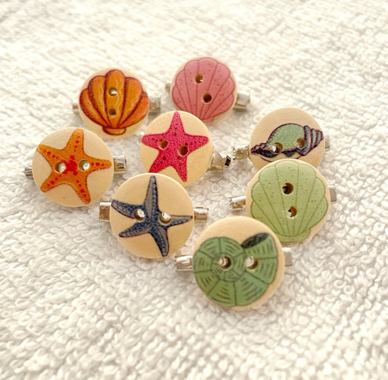 A set of eight wooden buttons with nautical designs, including starfish, seashells, and a pink starfish, arranged on a textured white fabric background.