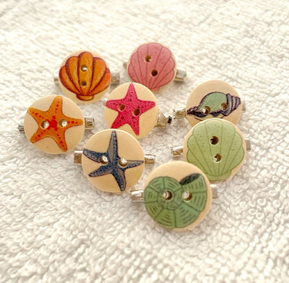 A set of eight wooden buttons with nautical designs, including starfish, seashells, and a pink starfish, arranged on a textured white fabric background.