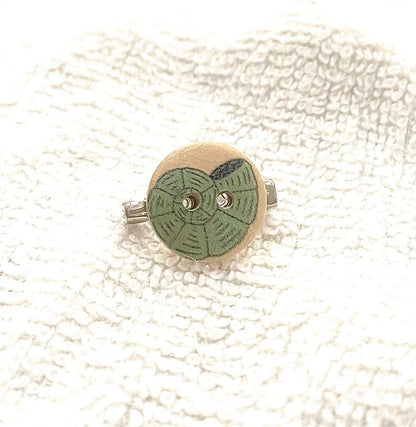 A small round wooden button with a green hand-painted geometric pattern and a silver metal loop, shown on a textured white fabric background.