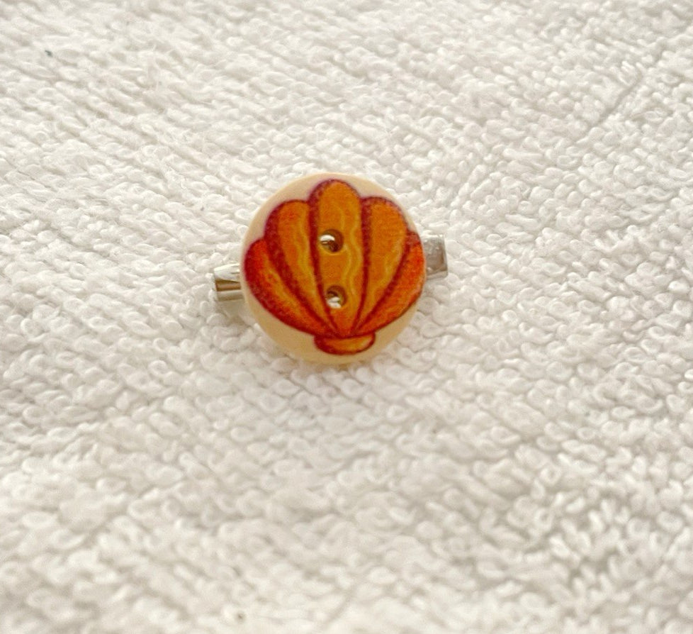 A small round button with a seashell design in orange, red, and yellow, mounted on a silver ring, displayed on a textured white fabric background.