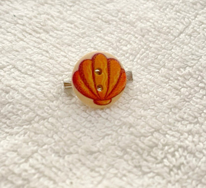 A small round button with a seashell design in orange, red, and yellow, mounted on a silver ring, displayed on a textured white fabric background.