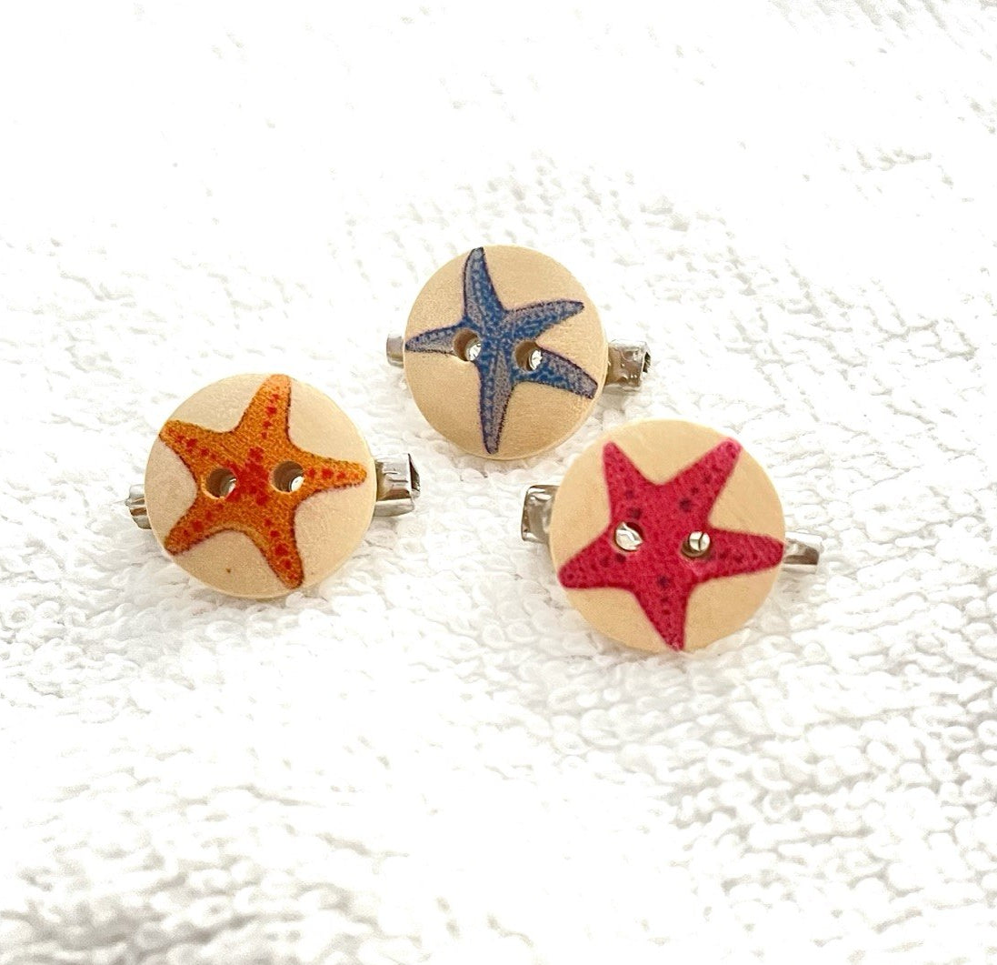 Three round wooden button brooches with starfish designs in orange, blue, and red, displayed on a textured white fabric background.