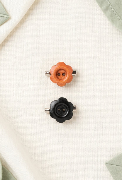 Two small flower-shaped metal buttons, one orange and one black, displayed on a textured cream fabric background.