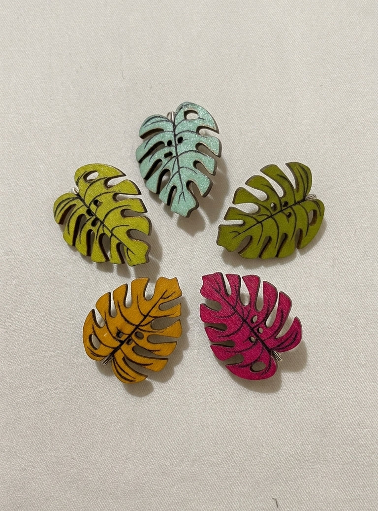 Five wooden monstera leaf-shaped brooches in yellow, mint green, olive green, mustard yellow, and magenta pink, arranged on a light beige textured fabric background.