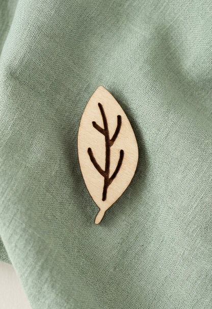 A small natural wood leaf-shaped brooch with a dark brown engraved leaf vein design, resting on a textured sage green fabric background.